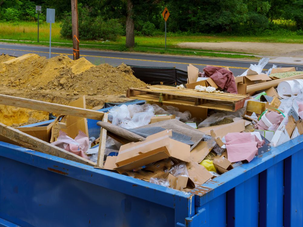 A cluttered dumpster on the side of the road, brimming with construction debris and litter.